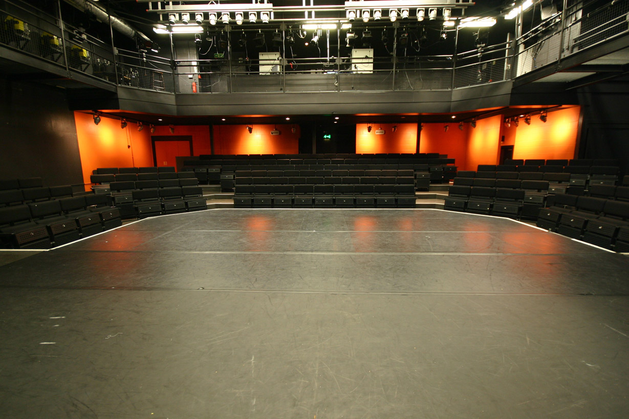 The Studio Theatre pictured from the stage. The black floor of the stage stretches away from the viewer, beyond it are rows of black upholstered seats and at the back of the room lights shine on an orange wall.