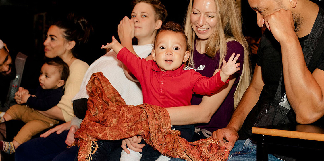 Smiling parents hold their babies in the audience for Bring Your Own Baby Comedy. In the centre of the image, a mother is grinning down at her baby, who looks straight to the camera with wide eyes and arms spread open!
