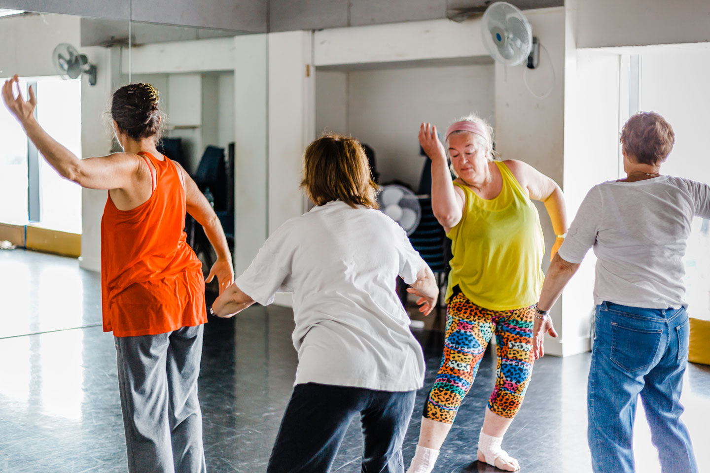 A group of four women dancing in the dance space. They are all in a different position, two wear brightly coloured sportswear.