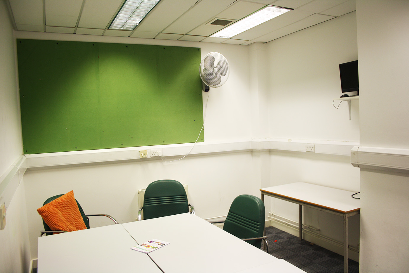 The artsdepot small meeting room set up for a meeting. There is space for multiple tables and chairs. On the back wall is a green noticeboard and a fan.
