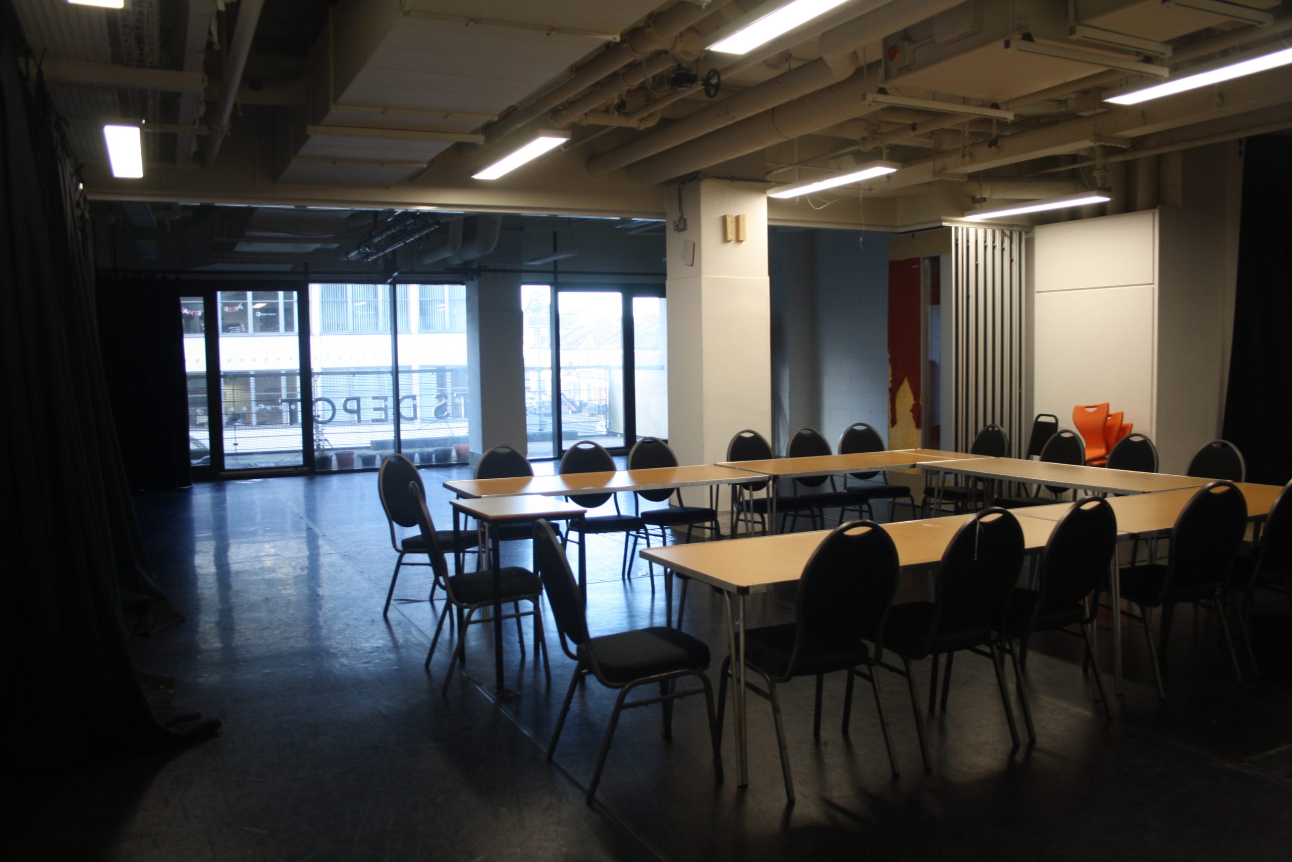 artsdepot creation space photographed from the entrance. The space is a large drama studio with black flooring and window panelling along the back wall to let in natural life. In the photo, a large U-shaped meeting table is assembled in the foreground of the image.