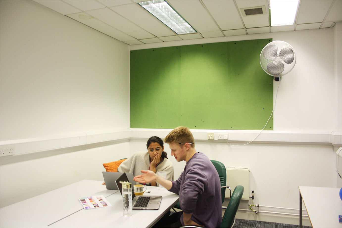 Two artsdepot team members converse in the meeting room. They are engaged in conversation with laptops open and water and coffee cups on the table. Behind them is a green noticeboard.