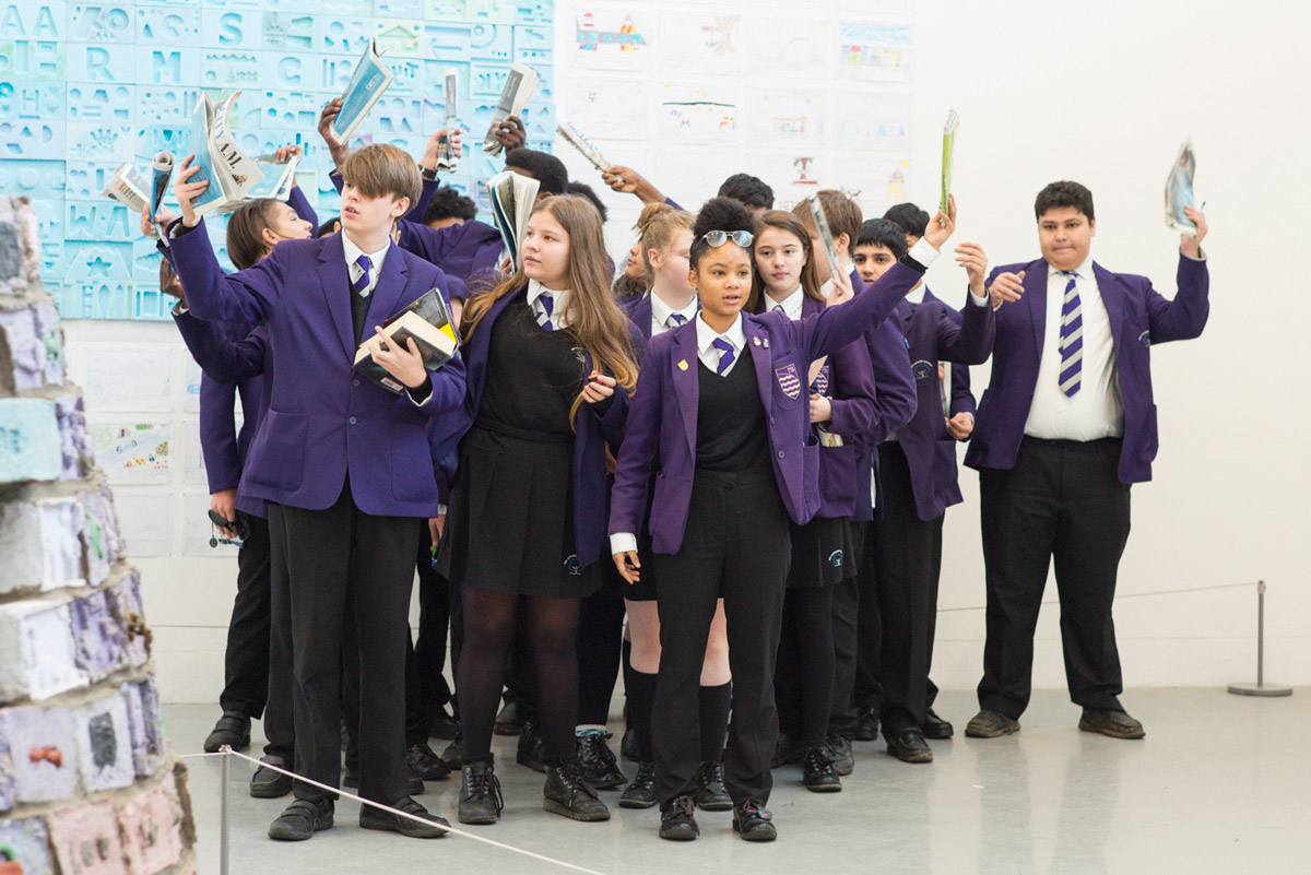 A group of students in purple school uniforms performing in a white-walled gallery. Some hold books, others hold newspapers in the air.