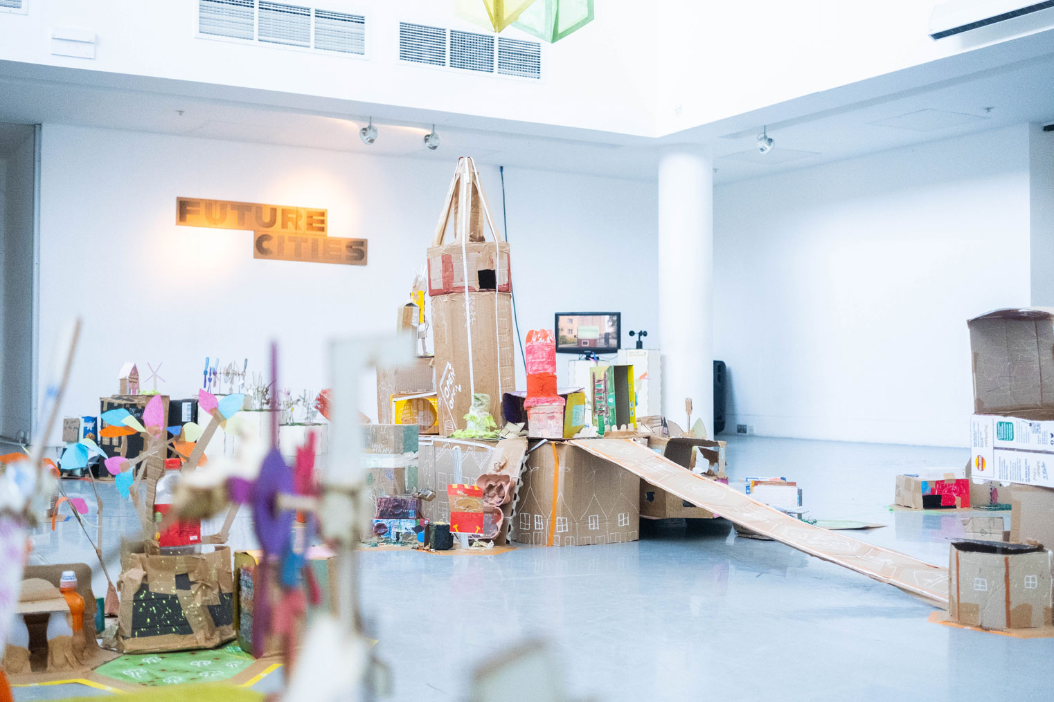 Structures made from cardboard, coloured paper and recycled materials on display in a white-walled gallery.