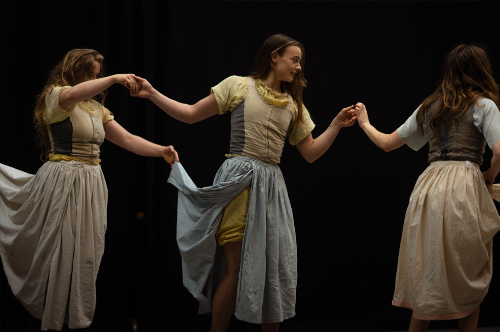 Rehearsal photography. Three female performers link hands, facing right, in movement. They wear rustic-style historical skirts and dresses.