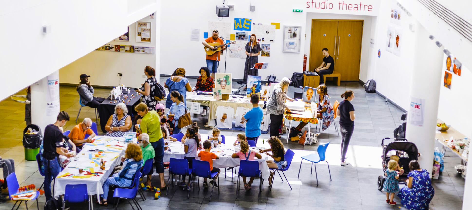 The artsdepot cafe area photographed in the middle of a busy event. Colourful chairs are filled with people of all ages, chatting and engaged with crafts. In the background, two musicians are performing, playing guitar and singing live.