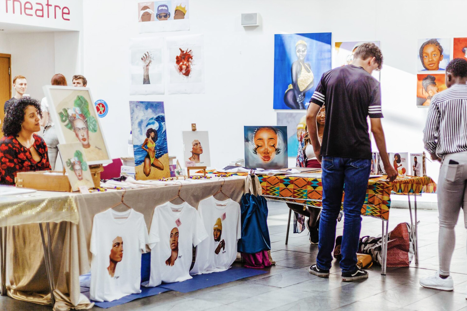 An artist displays her work at an arts and crafts showcase event. A customer is viewing her on-sale work. Her work features dynamic depictions of black womanhood in vibrant portraiture.