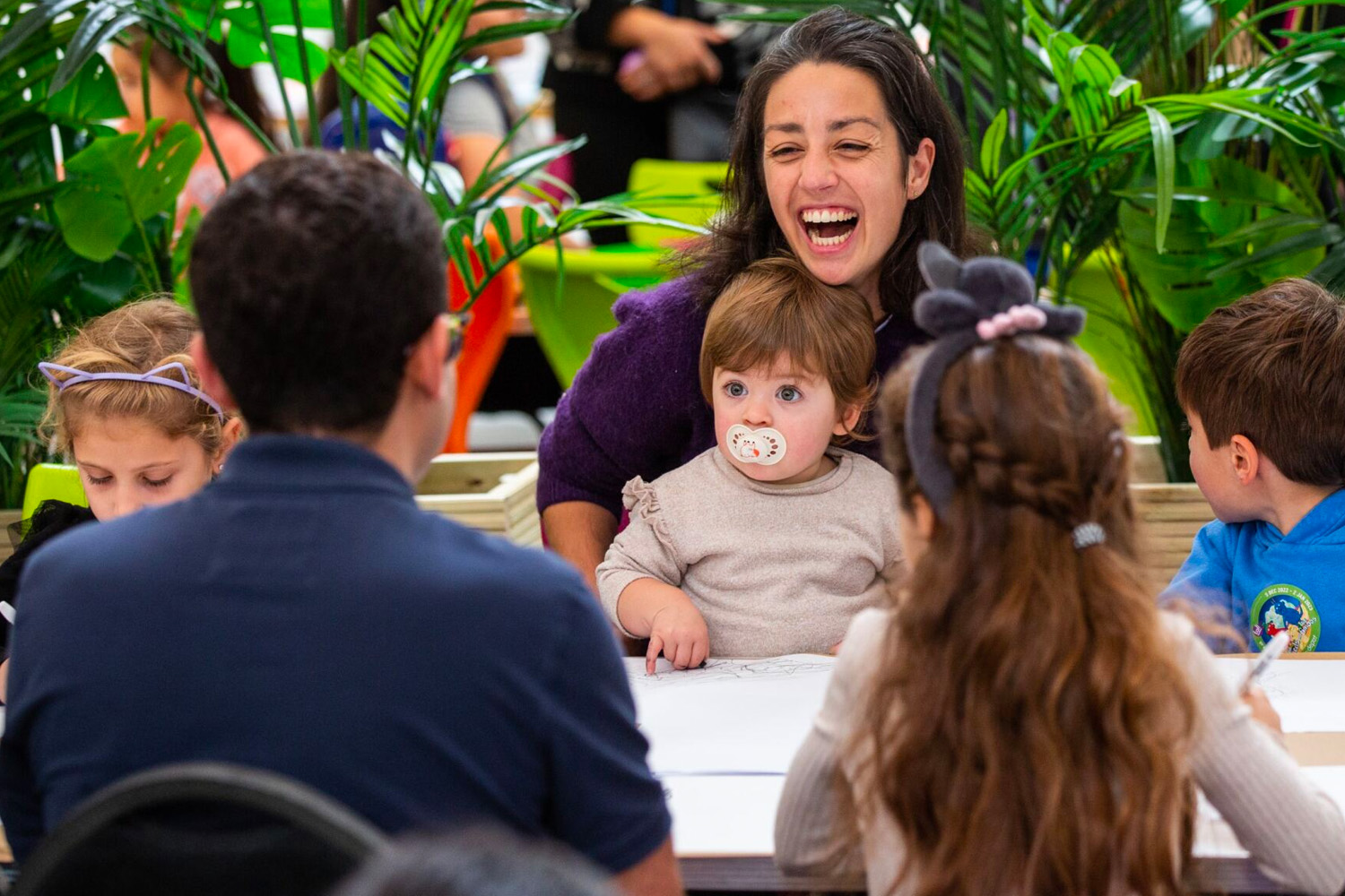 A family of six sit in the artsdepot cafe. In the centre sits mum, who is laughing with a big smile, holding her baby on her lap.