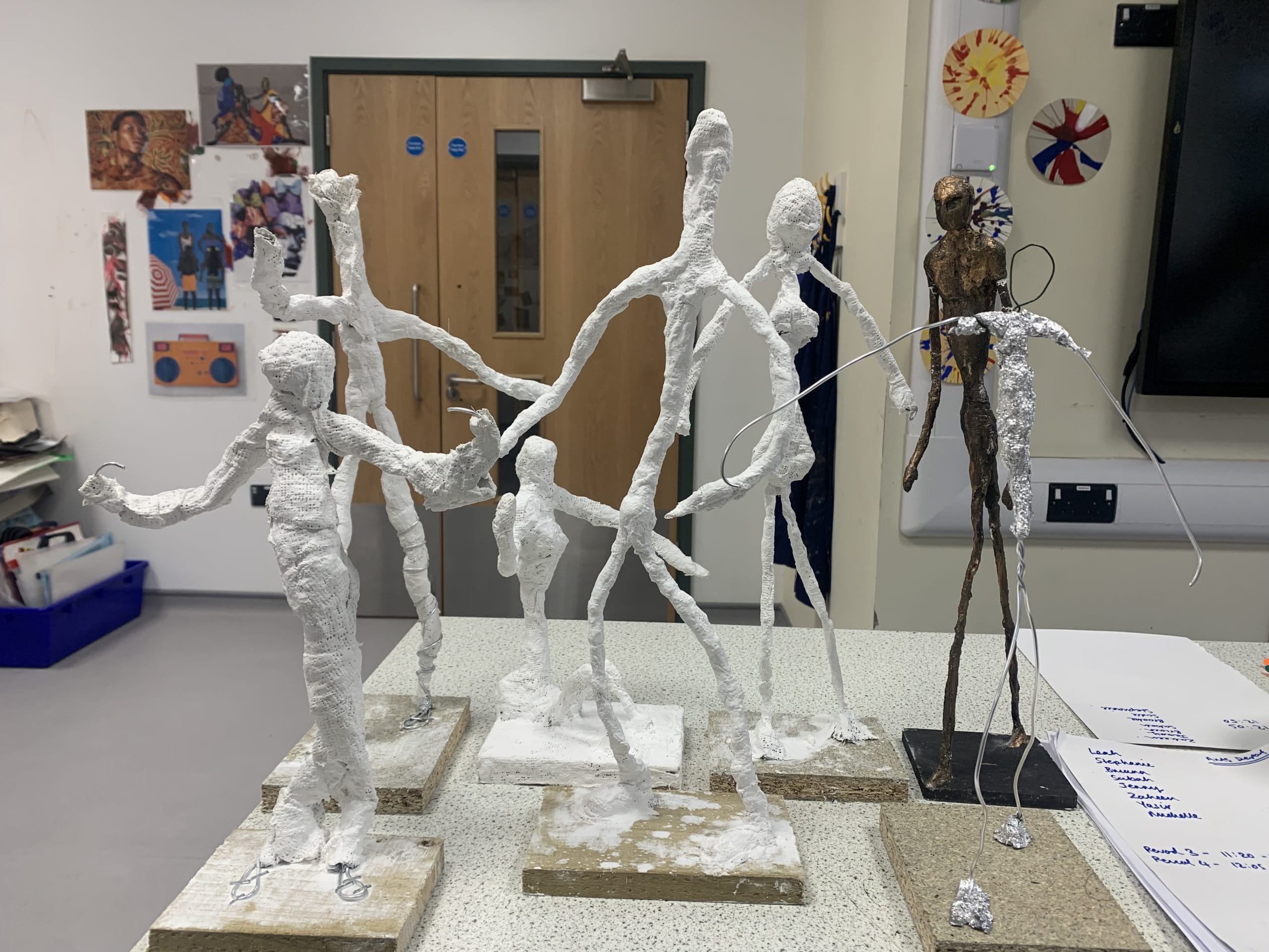 Several Papier-mâché figurines are posed on a table in an art studio. Some of the figurines are fully coated in Papier-mâché, whilst others are still wire frames. Colourful artwork is hanging in the background.
